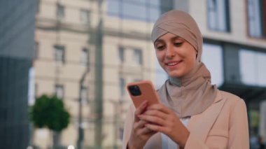 İş merkezinin yakınında akıllı telefon kullanarak duran tesettürlü güzel, genç, gülümseyen Arap iş kadını. Kapatın. İnsanlar ve teknoloji sektörü kavramı