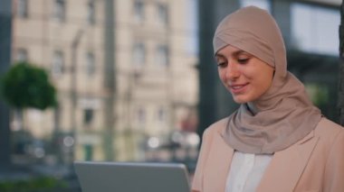 Mutlu Arap iş kadını tesettür takıyor, dışarıda çalışıyor, iş için dizüstü bilgisayar kullanıyor. Başörtülü Arap kadın iş merkezinin arka planında dizüstü bilgisayarla çalışıyor.