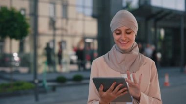 Mutlu Arap iş kadını tesettürlü, açık havada çalışıyor, tablet kullanıyor, sosyal medyayı araklıyor. İş merkezinin arka planında tablet üzerinde çalışan genç Arap bayan.