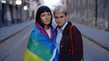 Ciddi bir lezbiyen çiftin portresi lgbt bayrağıyla kameraya bakıyor. Şehir merkezinde gökkuşağı bayrağıyla poz veren iki mutlu bayan arkadaş. Kadın ilişkileri, lgbt kavramı