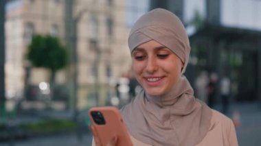 Güzel, genç, gülümseyen Arap iş kadını resmi kıyafetlerle tesettüre girmiş akıllı telefon kullanarak iş merkezinin yakınında yürüyor. Kapatın. İnsanlar ve teknoloji sektörü kavramı