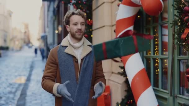 白人の幸せな男はクリスマスギフトパッケージをキャッチし カメラに笑顔と装飾されたクリスマスの背景にカメラを見てそれを示しています 人々とクリスマス休暇の概念 — ストック動画