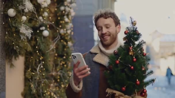 スマホアプリでクリスマスツリーを開催しているクリスマスツリーショップから帰ってきた幸せな白人男性 新しい年末年始 テクノロジーの概念 — ストック動画