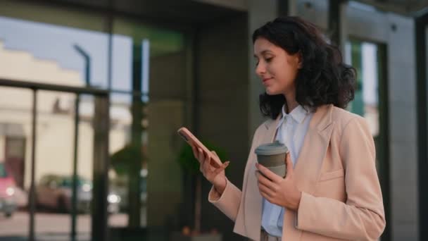 这位忙碌的 面带微笑的女商人一边拿着咖啡一边用智能手机走进办公大楼 一边给她的客户回发短信 人员和技术业务概念 — 图库视频影像