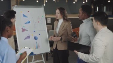 Kafkasyalı ciddi iş kadını finansal raporunu çizelgelerle sunuyor ve grafikleri ters yüz ediyor. Çok kültürlü bir ekip dikkatle dinliyor ve not alıyor..