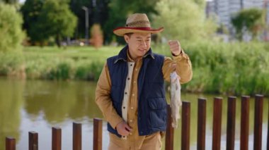 Özel balıkçı kıyafeti giymiş Asyalı bir adam oltaya takılmış balığı tutuyor ve kameraya bakarken mutlu oluyor.