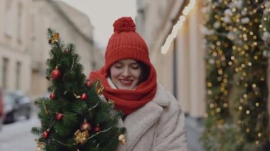 Mutlu gülümseyen beyaz kadın kışın dekore edilmiş caddede yürüyor Noel havasının tadını çıkarıyor kırmızı şapka ambalajıyla ağacı tutuyor. İnsanlar ve Noel tatili konsepti