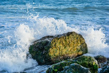 Büyük taşlı deniz kıyısı, deniz dalgaları, beyaz köpük, mavi su, güneş ışığı.
