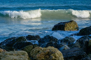 Büyük taşlı deniz kıyısı, deniz dalgaları, beyaz köpük, mavi su, güneş ışığı.