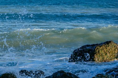 Büyük taşlı deniz kıyısı, deniz dalgaları, beyaz köpük, mavi su, güneş ışığı.