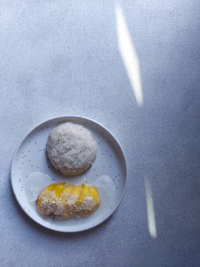 Bir tabak mango yapışkan pirincin seçici odağı. Güneşin gölgesi tabaklara düşüyor ve arka planda geleneksel Tayland sokak yemekleri var. Hindistan cevizi sütü ve susam tohumuyla tamamlandı..
