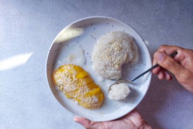 Bir tabak mango yapışkan pirincin seçici odağı. Güneşin gölgesi tabaklara düşüyor ve arka planda geleneksel Tayland sokak yemekleri var. Hindistan cevizi sütü ve susam tohumuyla tamamlandı..