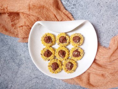 Çikolatalı tart kurabiyeleri. Beyaz tabakta servis ediliyor. un, yumurta sarısı, margarin, mısır nişastası, tereyağı, süt, arıtılmış şeker, tuz, çikolata parçacıkları ve vanilya özünden yapılır. Çikolata ezmesiyle süslenmiş.