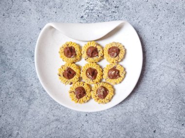 Çikolatalı tart kurabiyeleri. Beyaz tabakta servis ediliyor. un, yumurta sarısı, margarin, mısır nişastası, tereyağı, süt, arıtılmış şeker, tuz, çikolata parçacıkları ve vanilya özünden yapılır. Çikolata ezmesiyle süslenmiş.