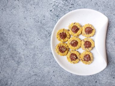 Çikolatalı tart kurabiyeleri. Beyaz tabakta servis ediliyor. un, yumurta sarısı, margarin, mısır nişastası, tereyağı, süt, arıtılmış şeker, tuz, çikolata parçacıkları ve vanilya özünden yapılır. Çikolata ezmesiyle süslenmiş.