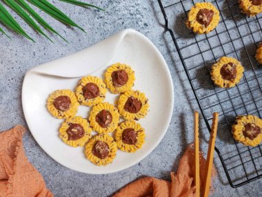 Çikolatalı tart kurabiyeleri. Beyaz tabakta servis ediliyor. un, yumurta sarısı, margarin, mısır nişastası, tereyağı, süt, arıtılmış şeker, tuz, çikolata parçacıkları ve vanilya özünden yapılır. Çikolata ezmesiyle süslenmiş.