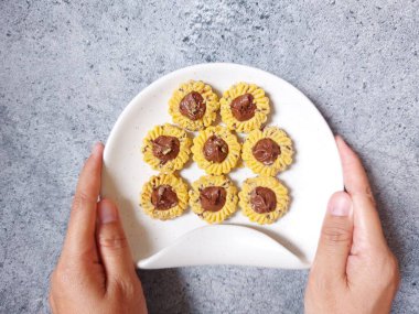 Çikolatalı tart kurabiyeleri. Beyaz tabakta servis ediliyor. un, yumurta sarısı, margarin, mısır nişastası, tereyağı, süt, arıtılmış şeker, tuz, çikolata parçacıkları ve vanilya özünden yapılır. Çikolata ezmesiyle süslenmiş.