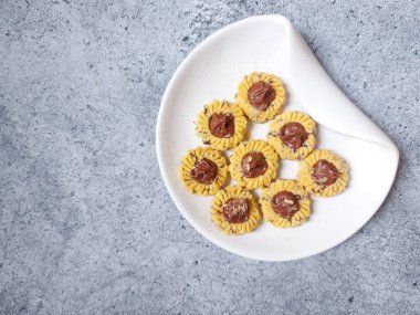 Çikolatalı tart kurabiyeleri. Beyaz tabakta servis ediliyor. un, yumurta sarısı, margarin, mısır nişastası, tereyağı, süt, arıtılmış şeker, tuz, çikolata parçacıkları ve vanilya özünden yapılır. Çikolata ezmesiyle süslenmiş.