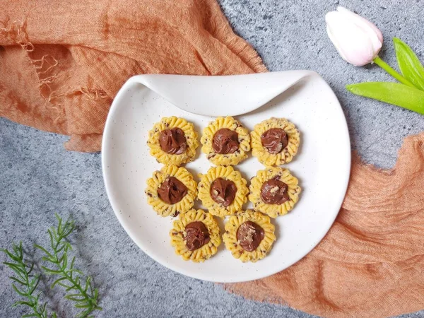 Çikolatalı tart kurabiyeleri. Beyaz tabakta servis ediliyor. un, yumurta sarısı, margarin, mısır nişastası, tereyağı, süt, arıtılmış şeker, tuz, çikolata parçacıkları ve vanilya özünden yapılır. Çikolata ezmesiyle süslenmiş.