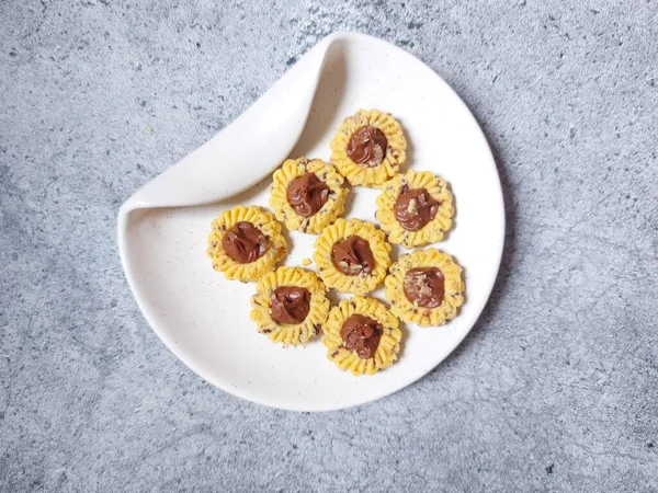 Çikolatalı tart kurabiyeleri. Beyaz tabakta servis ediliyor. un, yumurta sarısı, margarin, mısır nişastası, tereyağı, süt, arıtılmış şeker, tuz, çikolata parçacıkları ve vanilya özünden yapılır. Çikolata ezmesiyle süslenmiş.