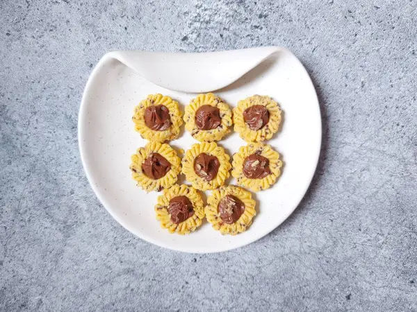 Çikolatalı tart kurabiyeleri. Beyaz tabakta servis ediliyor. un, yumurta sarısı, margarin, mısır nişastası, tereyağı, süt, arıtılmış şeker, tuz, çikolata parçacıkları ve vanilya özünden yapılır. Çikolata ezmesiyle süslenmiş.