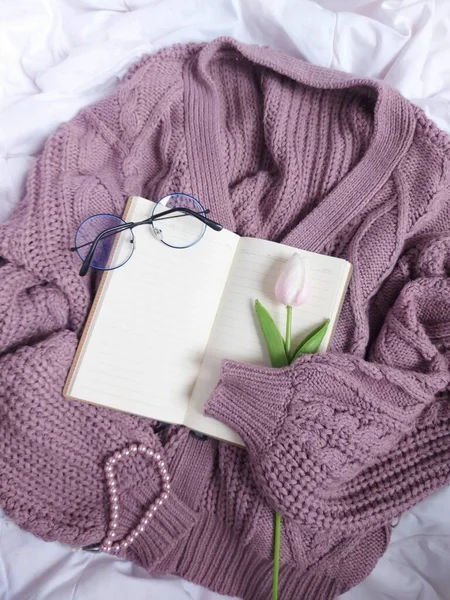A blank book agenda hold by purple sweater. Completed with glasses, pink tulip flower and pearl bracelet. Above white fabric. Still life photography concept.