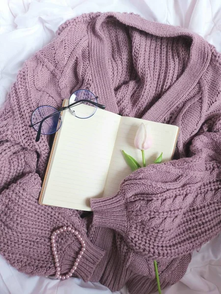 A blank book agenda hold by purple sweater. Completed with glasses, pink tulip flower and pearl bracelet. Above white fabric. Still life photography concept.