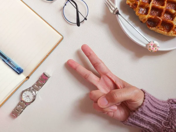 female hands forming peace symbol. completed with blank agenda book, glasses, knitted sweater, dessert, plate and fork. work break time photography concept