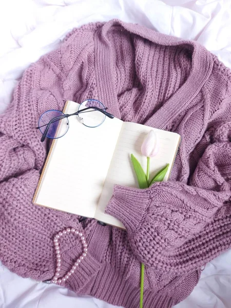 A blank book agenda hold by purple sweater. Completed with glasses, pink tulip flower and pearl bracelet. Above white fabric. Still life photography concept.