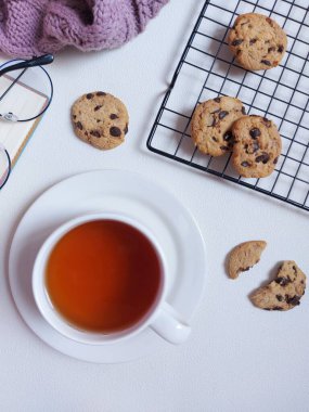 Tea time photography concept. A cup of hot tea. Completed with choco chips cookies, white plate, cooling rack, glasses,book and purple knitted