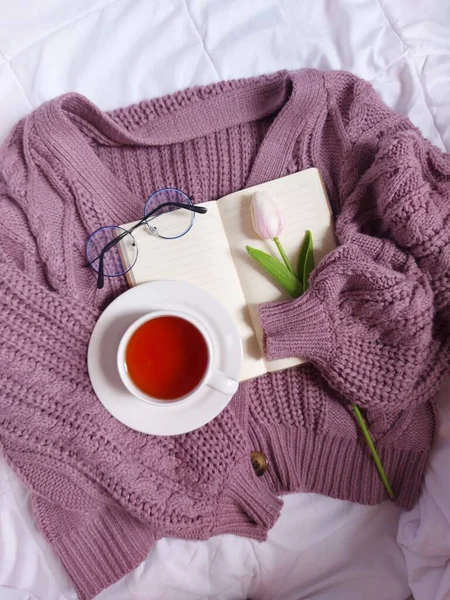 blank book agenda hold by purple sweater. Completed with glasses, pink tulip flower and a cup.of tea. Above white fabric. Still life photography concept.