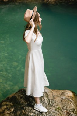 Beyaz uzun elbiseli ve şapkalı güzel bir kadın suyun kenarında duruyor ve gülümsüyor. Seyahat kavramı.