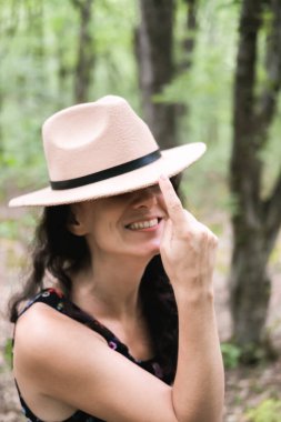 Genç milenyum kadını, elbiseli ve bej şapkalı ormanda mutlu ve rahat duruyor, doğanın ve huzurun tadını çıkarıyor. Yalnız seyahat konsepti.