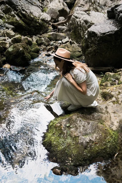 身穿白色长裙、头戴白色帽子的漂亮女人坐在河边笑着。旅行概念. — 图库照片