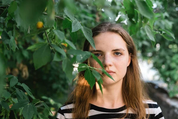 Retrato de jovem linda mulher com olhos azuis e sardas em camiseta listrada e jeans contra fundo de folhagem verde. Fotografias De Stock Royalty-Free