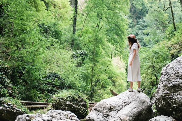 Mulher jovem milenar em vestido e chapéu bege fica na floresta feliz e relaxado, goza de natureza e tranquilidade. Viajar sozinho conceito. Fotografia De Stock