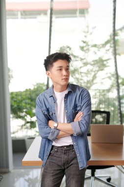 Modern ofiste dururken kollarını kavuşturan neşeli Asyalı genç adam.