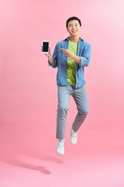 興奮した若い男は空中でジャンプ ピンクの背景に黒の空の画面を持つ携帯電話を示します — ストック写真