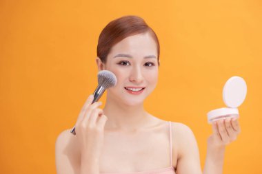 Güzel bir kadın büyük bir kozmetik fırçasıyla yanağına allık sürüyor. Önünde küçük bir ayna tutuyor.