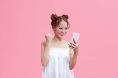 Cheerful asian woman standing isolated over pink background, holding mobile phone, celebrating