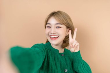 Gülümseyen sevimli Asyalı kadın, günlük kıyafetlerle fotoğraf çekiyor ve bej arka plandan kameraya bakıyor.