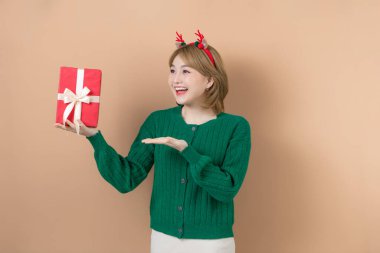 Eğlenceli dekoratif geyik boynuzları takmış genç bir kız bej arka planda izole edilmiş hediyesiyle birlikte.