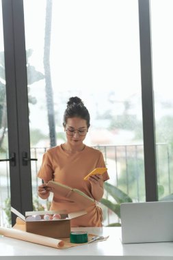 Güzel bayan girişimci cep telefonundan siparişleri kontrol ederken paket kutuları ve eşyaları hazırlıyor.