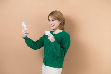 İnternetten alışveriş. Bej arka planda cep telefonu ve banka kartıyla duran gülümseyen kadın portresi