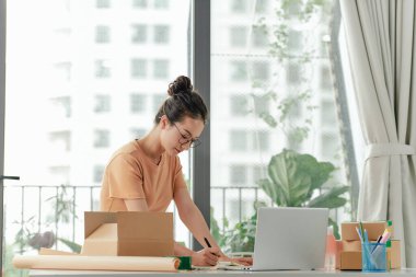 Kadın dizüstü bilgisayarda siparişleri kontrol ediyor ve teslimatı paket kutusuna yazıyor.