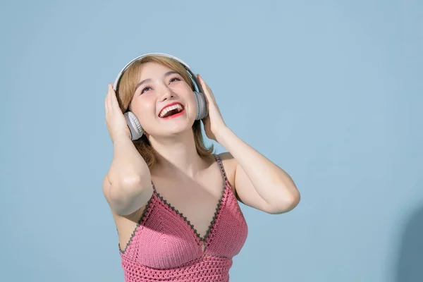 Studio shot of happy Asian millennial girl smiles gladfully looks away uses smartphone and headphone