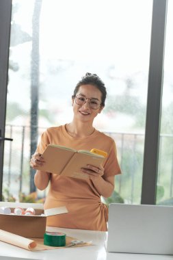 Güzel bayan girişimci cep telefonundan siparişleri kontrol ederken paket kutuları ve eşyaları hazırlıyor.