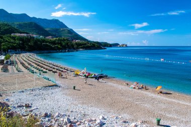 Rafailovici, Karadağ - 3 Temmuz 2021: Kamenovo çakıl taşı plajında güneşlik ve şemsiyeli tatilciler. Yaz güneşli bir sabah. Mavi gökyüzü. Sveti Stefan adasının arka planında. Budva Riviera.