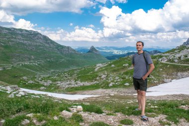 Güzel dağ zirvelerinin arka planındaki erkek turist, karlı yeşil tepeler ve bulutlu mavi gökyüzü. Durmitor Ulusal Parkı. Karadağ 'daki en yüksek yol geçidi. Güzel yaz manzarası.