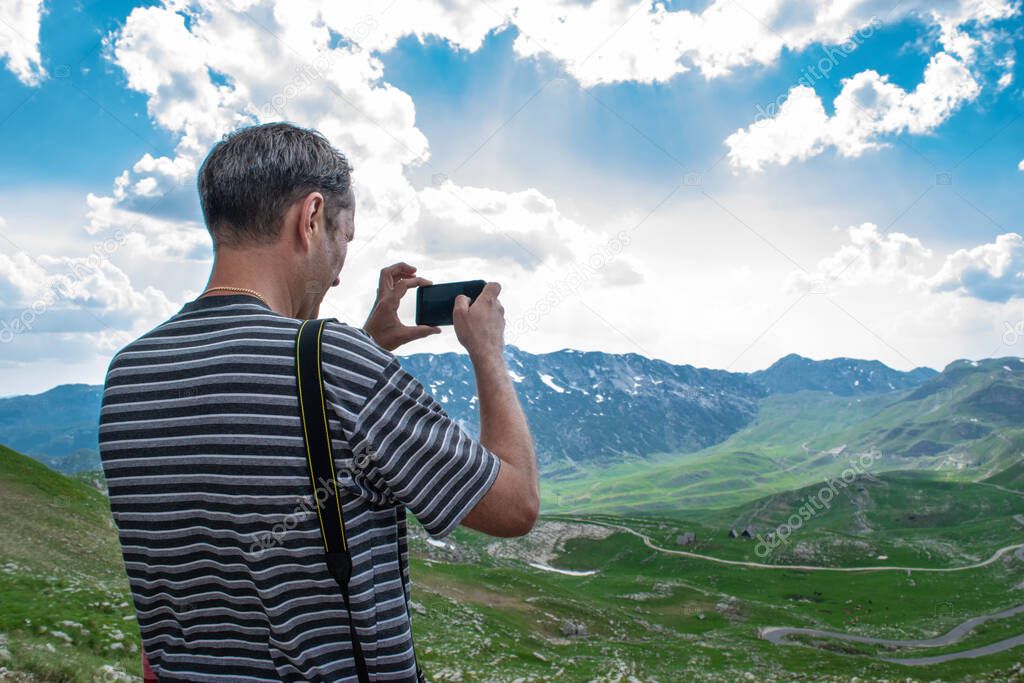 Man tourist takes a photo of beautiful summer landscape. Mountain peaks, green hills and cloudy blue sky of Durmitor National Park. Highest road pass in Montenegro.
